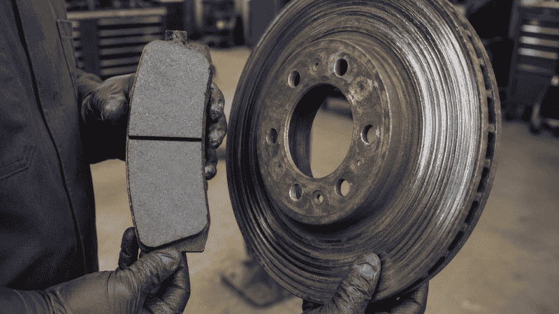 A mechanic holding a new brake pad next to a damaged worn rotor in an auto repair shop, illustrating the cost difference between timely and delayed brake repairs