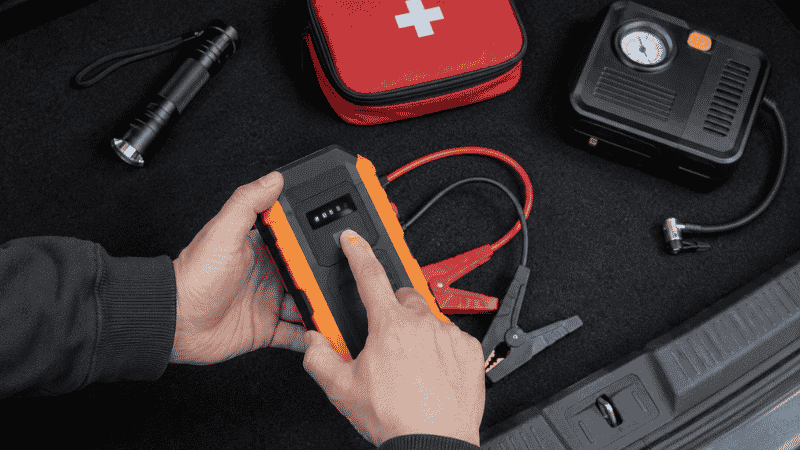 Person checking a portable jump starter charge level while reviewing car emergency items laid out on a trunk