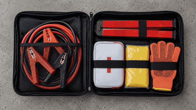 An open roadside emergency kit bag showing jumper cables, reflective triangles, first aid kit, rain poncho, and work gloves