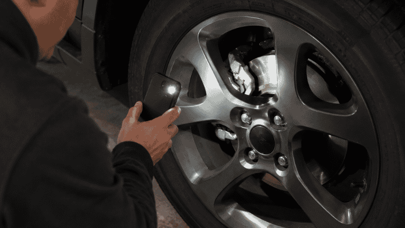 A person shining a phone flashlight through car wheel spokes to inspect the brake pad thickness against the rotor