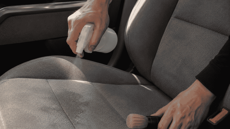 Spraying upholstery cleaner on a gray fabric car seat and scrubbing with a soft brush