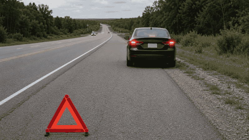A car pulled over on a wide road shoulder with hazard lights flashing and a reflective triangle placed behind it on the road