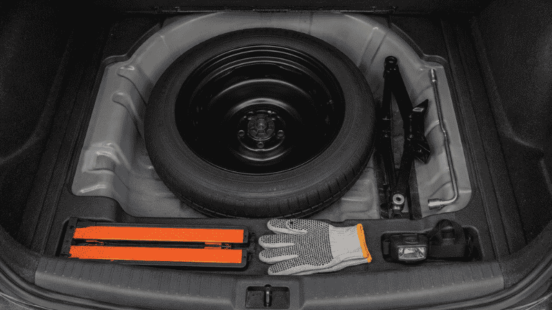Flat tire emergency tools laid out in a car trunk: scissor jack, lug wrench, spare tire, reflective triangles, gloves, and a flashlight