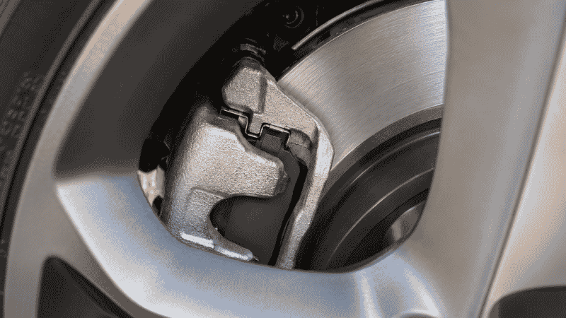 Close-up of a car brake disc and caliper through a wheel spoke, showing the brake pad pressed against the rotor