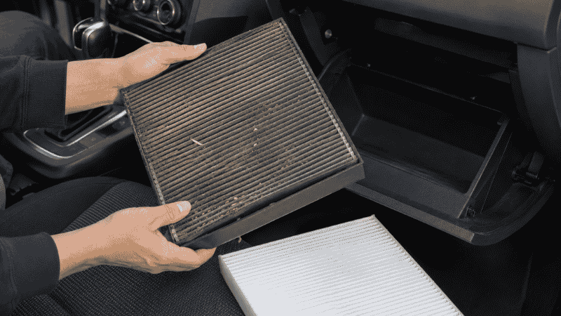 Hands removing a dirty cabin air filter from behind a car glove box to replace it