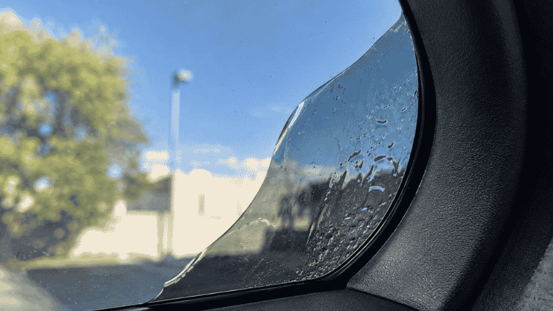 A car window showing bubbling and peeling window tint film in the corner, caused by repeated use of ammonia-based glass cleaner on tinted windows