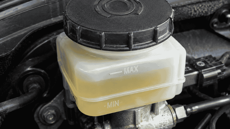 A car brake fluid reservoir near the firewall showing clear yellow brake fluid between the MIN and MAX lines, with the brake symbol visible on the cap