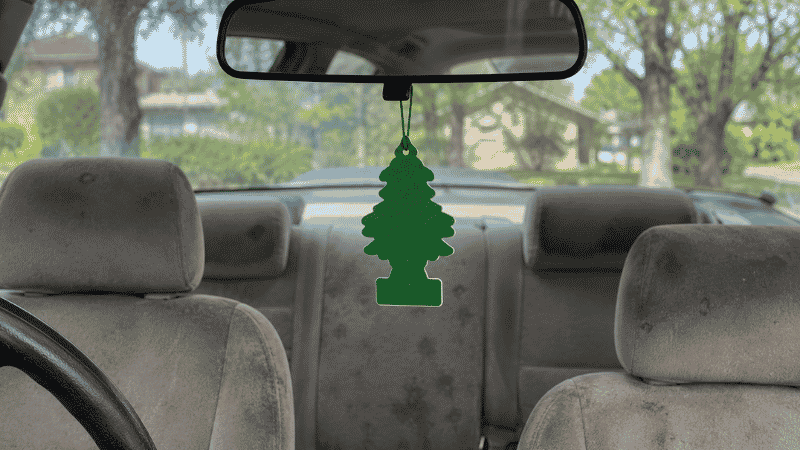 A pine tree shaped car air freshener hanging from a rearview mirror inside a car with visibly stained fabric seats, illustrating that fresheners mask but don't eliminate odors
