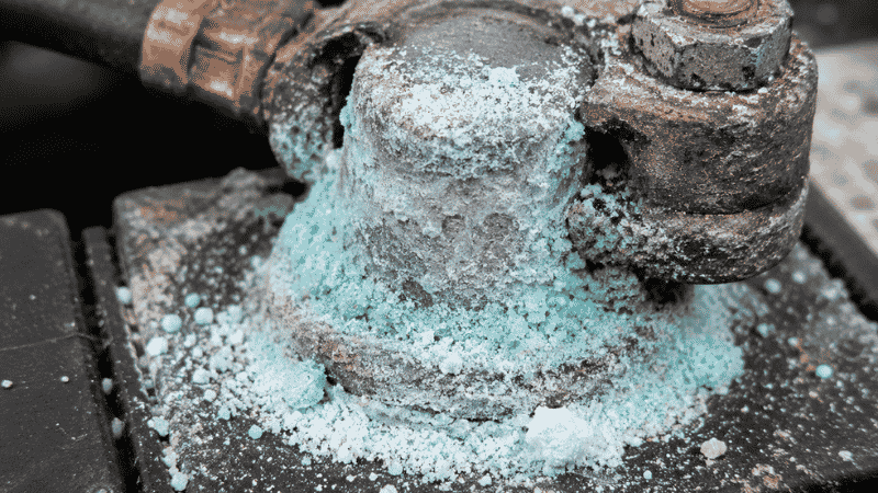 Close-up of a car battery terminal showing heavy white and blue corrosion buildup around the post and cable clamp connection