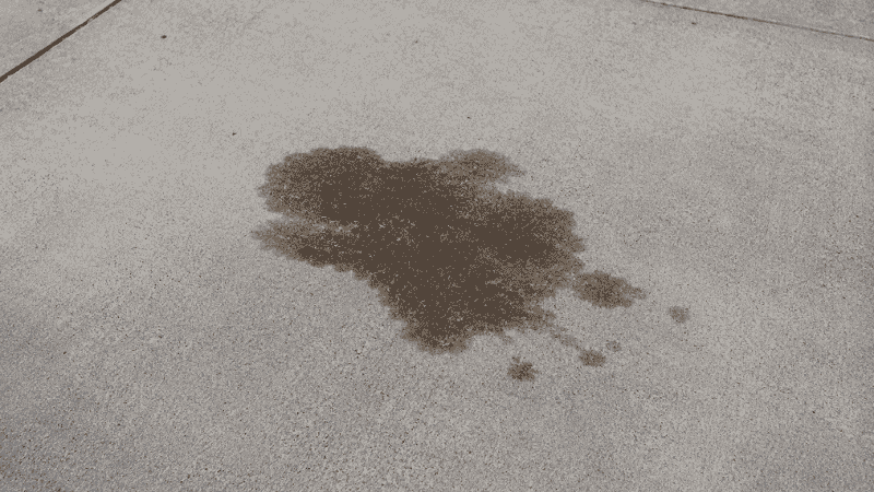 A dark oil stain puddle on a concrete driveway under where a car was parked, indicating an engine oil leak