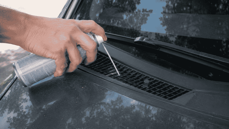 A person spraying HVAC duct cleaner into the fresh air intake vent at the base of a car windshield while the engine runs to eliminate odors from the ventilation system