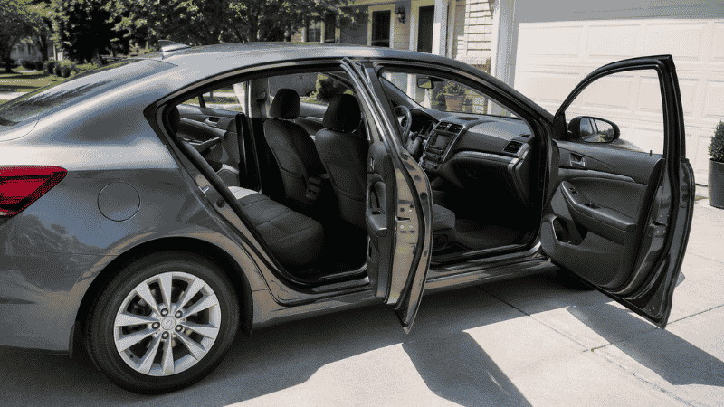 A car with all four doors open and windows down in a sunny driveway, allowing freshly cleaned car seats to air dry completely