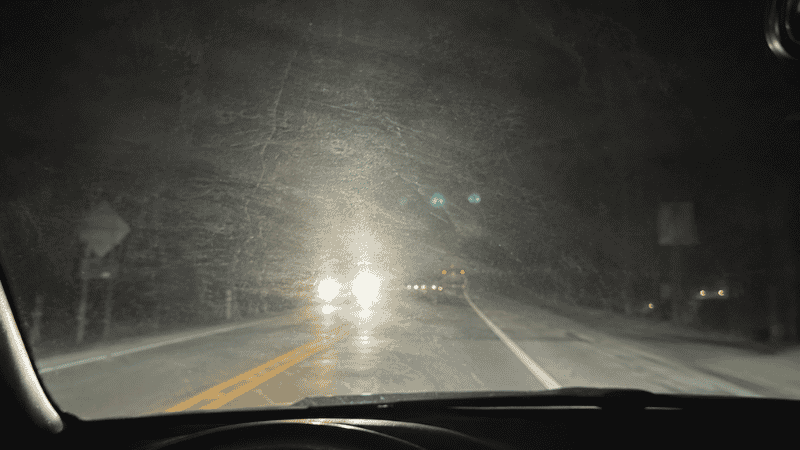 Driver's view through a hazy car windshield at night showing oncoming headlight glare and reduced visibility caused by the oily film on the interior glass