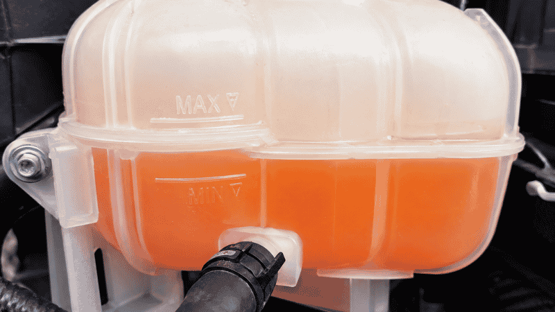 A translucent white plastic coolant reservoir in a car engine bay showing the orange coolant level between the MIN and MAX markings on the side