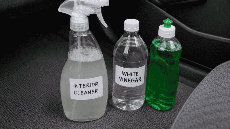 A spray bottle with homemade cleaning solution next to white vinegar and dish soap, ready to clean fabric car seats