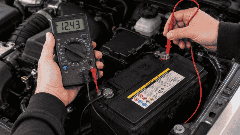 A person using a digital multimeter to test a car battery voltage in an engine bay, with the red and black probes connected to the battery terminals