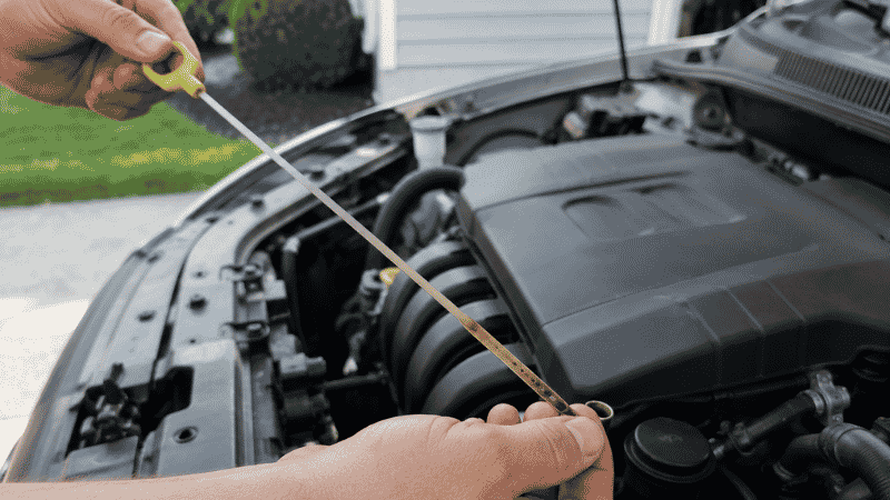 A person checking engine oil level with a dipstick under the open hood of a car in a residential driveway