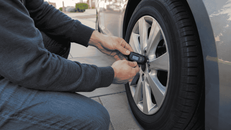 A person crouching beside a car tire pressing a digital tire pressure gauge onto the valve stem to check the tire pressure