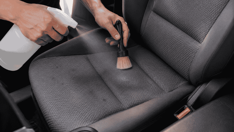 A person spraying upholstery cleaner on a gray fabric car seat and scrubbing with a soft brush to remove stains