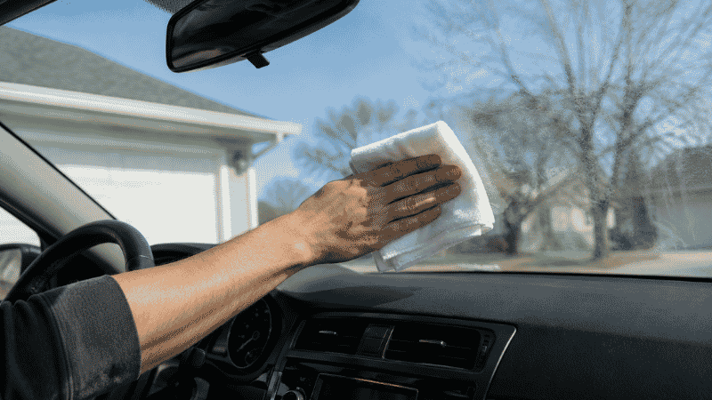 A person wiping a car windshield with a white microfiber cloth to achieve a streak-free clean on the interior glass