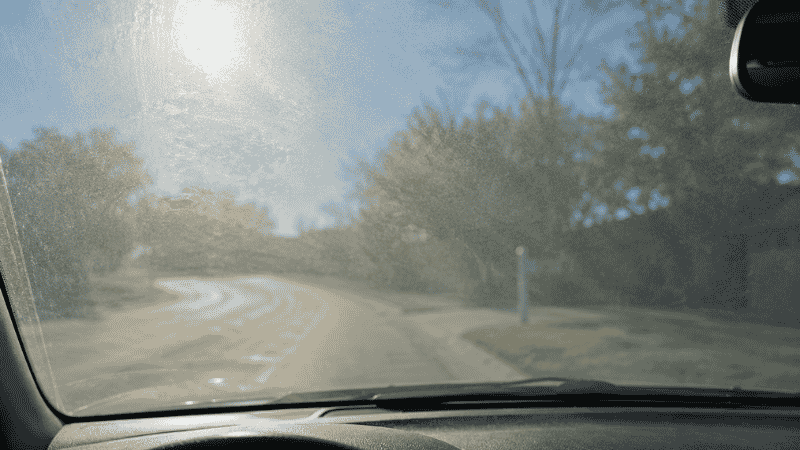 A car interior windshield photographed from the driver's seat showing the characteristic hazy oily film that develops on the inside of the glass from dashboard off-gassing