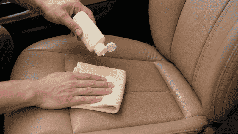 A person applying leather conditioner to a tan leather car seat with a microfiber cloth after cleaning, to restore moisture and prevent cracking