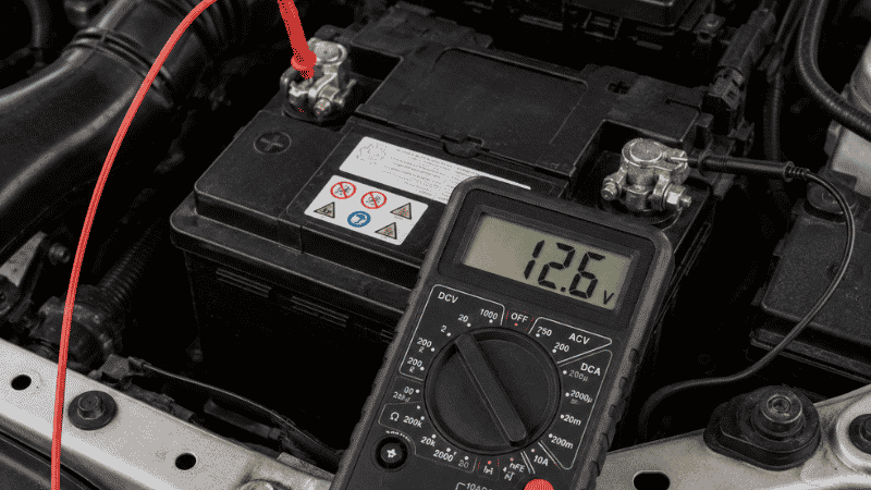 A digital multimeter displaying a 12.6 volt reading connected to a car battery with red probe on positive and black probe on negative terminal