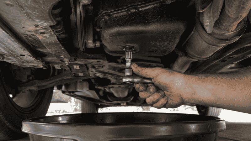A person's hand using a socket wrench to loosen the oil drain plug on the bottom of a car engine oil pan, with a black drain pan positioned below to catch the oil