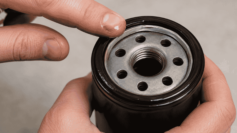 A person's finger applying a thin coat of fresh motor oil around the rubber gasket of a new oil filter before installation