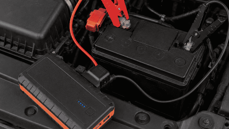 A compact lithium portable jump starter with red and black clamps connected to a car battery terminal in an engine bay, ready to start a dead battery