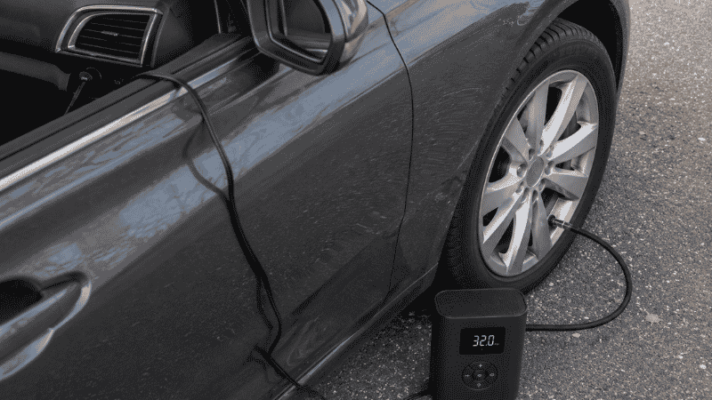 A compact 12V portable tire inflator connected to a car's power outlet with the hose attached to a tire valve stem, inflating the tire automatically