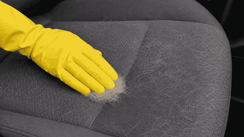 A hand wearing a rubber glove dragging across a car fabric seat to remove embedded pet hair, showing clumped fur being lifted from the surface