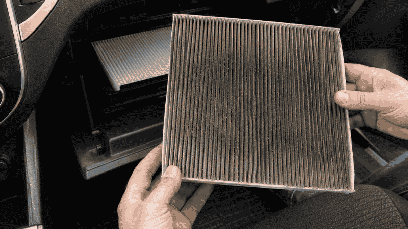 A dirty discolored cabin air filter being removed from behind a car glove box, showing the accumulated smoke particles and debris that cause interior odors