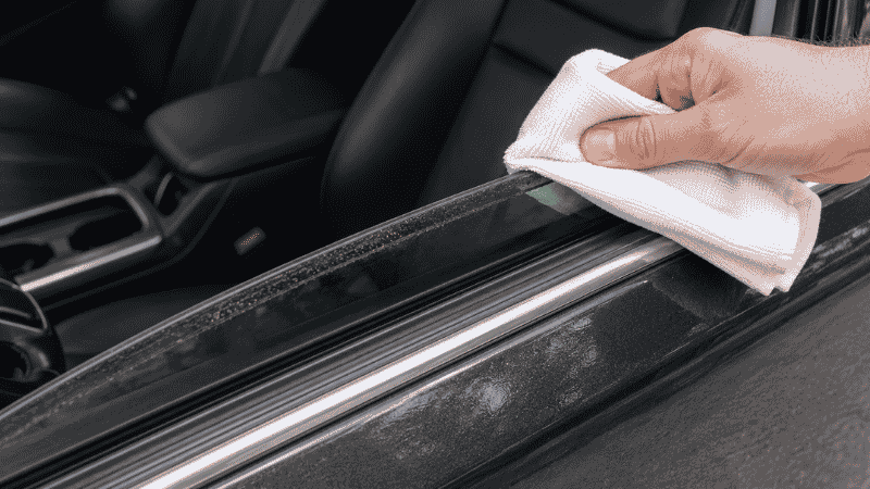 A car window rolled down one inch showing the dirty top edge of the glass that is normally hidden inside the door frame, being wiped clean with a microfiber cloth