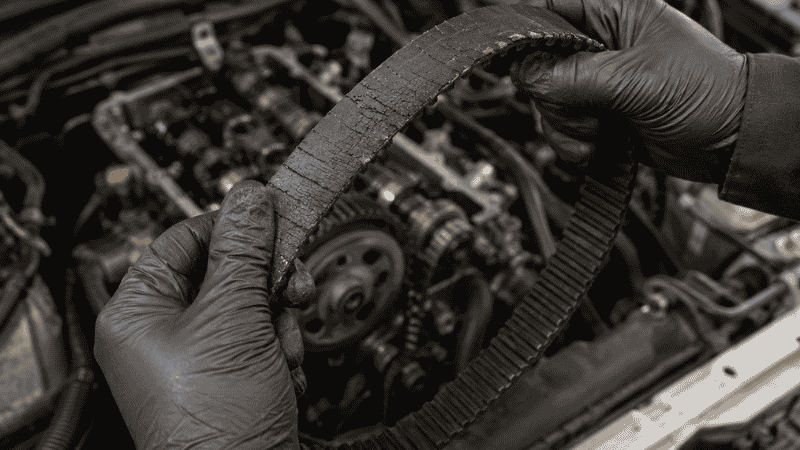 A mechanic's hands holding a worn rubber timing belt removed from a car engine, showing the importance of timely replacement before failure