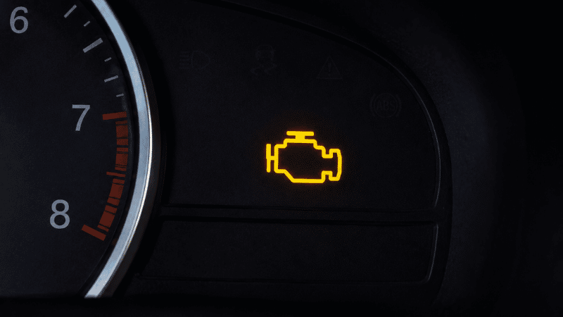 A car dashboard showing the check engine light illuminated in yellow-orange, representing the engine management warning symbol that requires diagnostic scanning