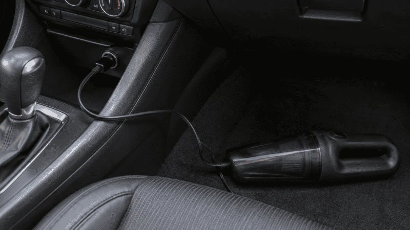 A 12V plug-in car vacuum connected to a car cigarette lighter power outlet showing the limited cord reach and power constraints of outlet-powered car vacuums
