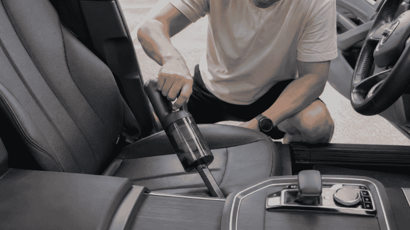 A person using a cordless handheld vacuum to clean a car seat gap and center console crevice during a quick interior cleaning session