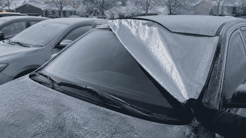 A car sun shade being removed from a windshield on a cold winter morning revealing a frost-free clear glass surface underneath while surrounding cars have frost-covered windshields