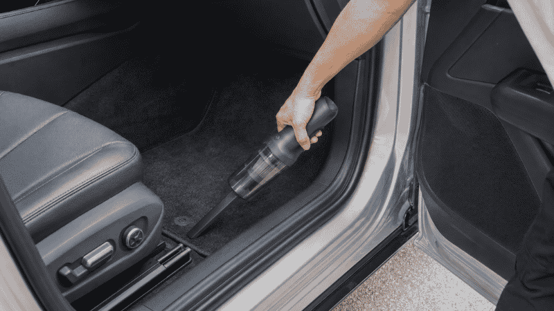A compact cordless handheld vacuum cleaner being used to vacuum a car floor mat, showing the practical size and maneuverability for car interior cleaning