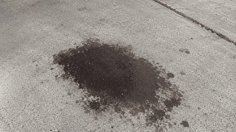 A dark brown engine oil stain puddle on a concrete driveway where a car was parked, indicating an oil leak that needs to be investigated when oil level keeps dropping