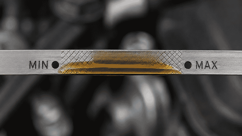 Close-up of an oil dipstick tip showing the oil level sitting between the MIN and MAX marks with amber-colored engine oil clearly visible on the metal stick