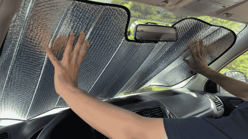A person installing a car sun shade on the windshield with the reflective silver side facing outward toward the sun, demonstrating correct installation orientation