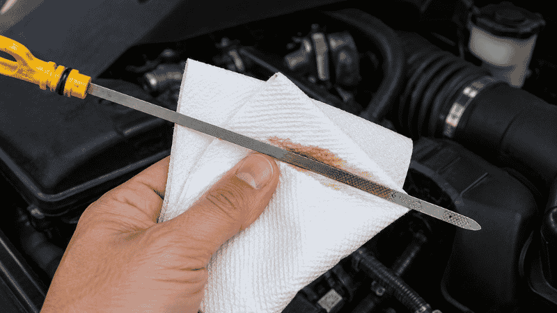 A person wiping an engine oil dipstick clean with a white paper towel from handle end toward the tip before reinserting it for an accurate oil level reading