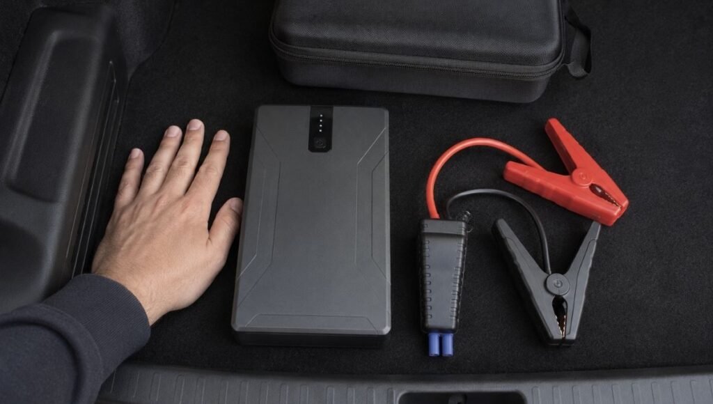 A compact lithium portable jump starter stored in a car trunk alongside its carrying case showing the small size relative to a hand for scale
